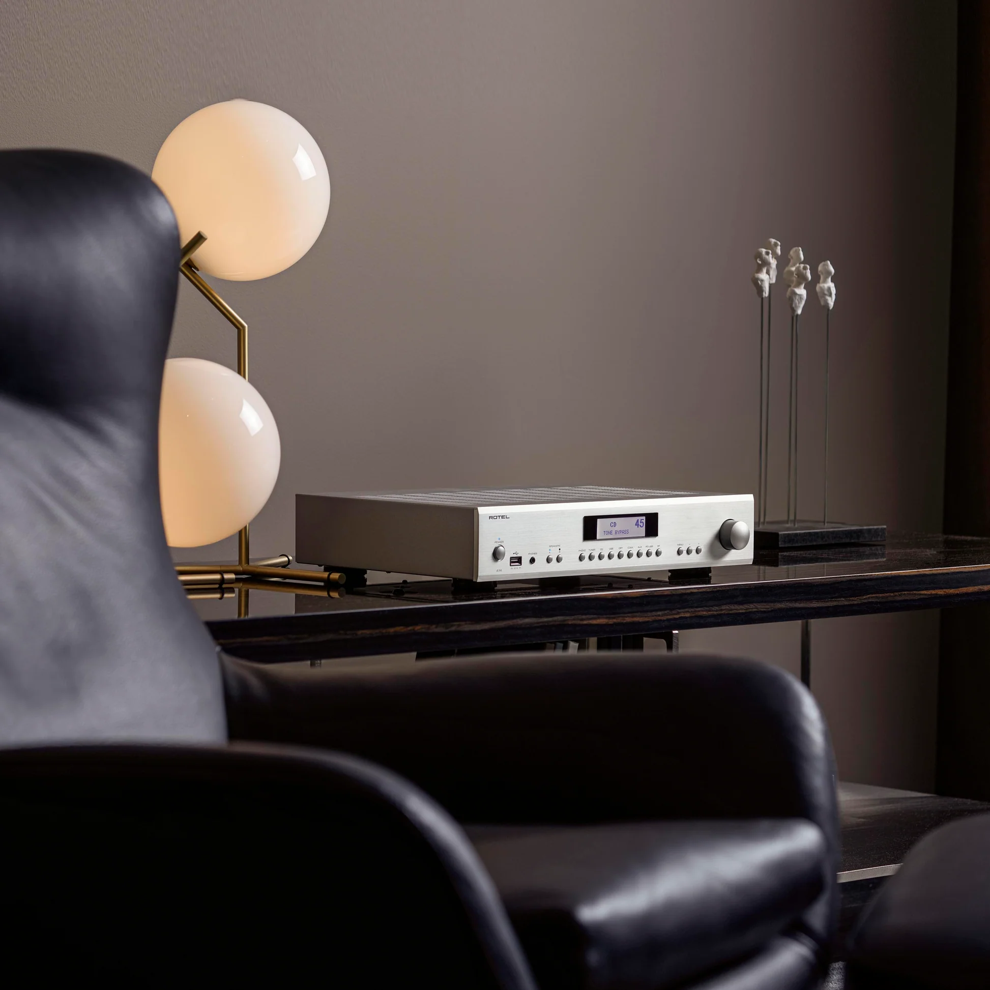 מגבר סטריאו משולב Rotel A14MkII בגימור כסף על שידת עץ כהה בסלון מעוצב, המציג חזית אלגנטית עם מסך LCD גרפי לצד גוף תאורה מודרני