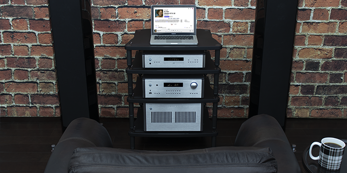 קדם-מגבר RC-1590MKII כסוף במרכז מעמד אודיו, בין נגן CD למגבר כוח. צג טקסט כחול ועיצוב אלגנטי על רקע קיר לבנים