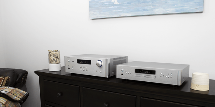 מגבר Rotel RA-1572MKII בכסף לצד נגן CD תואם על שידה שחורה. המגבר מציג צג דיגיטלי כחלחל וחוגת ווליום עם תאורה כחולה, על רקע קיר לבן עם תמונה