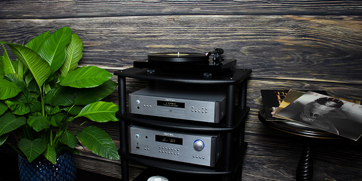 מגבר משולב Rotel RA-1592MKII בכסף על מעמד אודיו שחור, תחת נגן CD ופטיפון. המגבר מציג צג פלורסנט כחלחל, שורת כפתורי בחירת מקור וחוגת ווליום עם תאורה כחולה, על רקע קיר עץ כהה וצמחייה