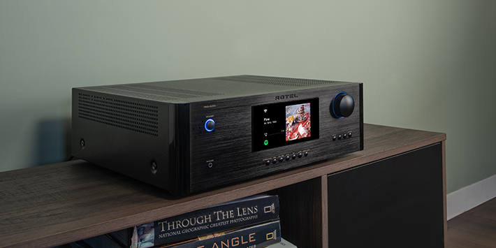 מגבר סטריאו וסטרימר Rotel RAS-5000 בשחור מוברש על שידת עץ. במרכז צג צבעוני המציג עטיפת אלבום, כפתור הפעלה וחוגת ווליום מוארים בכחול, על רקע קיר ירוק