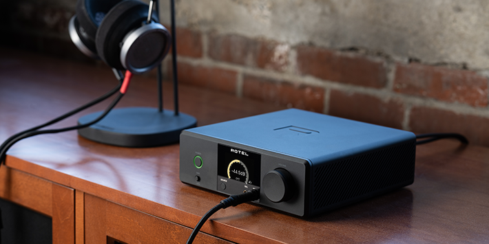 מגבר אוזניות ו-DAC דגם Rotel DX-3 בשחור על שידת עץ. המכשיר מציג עוצמה של -44.5dB על מסך OLED וכפתור הפעלה מואר בירוק, לצד אוזניות מקצועיות על רקע קיר לבנים