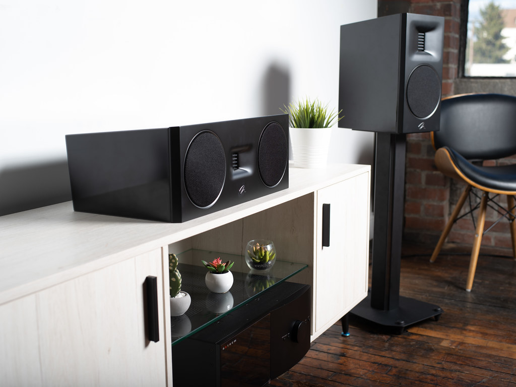 זוג רמקולים מרטין לוגן שחורים Martin Logan Motion XT B100 עם הסנטר התואם מונח על שידה לבנה ומשני צידיה הרמקולים בסלון מודרני