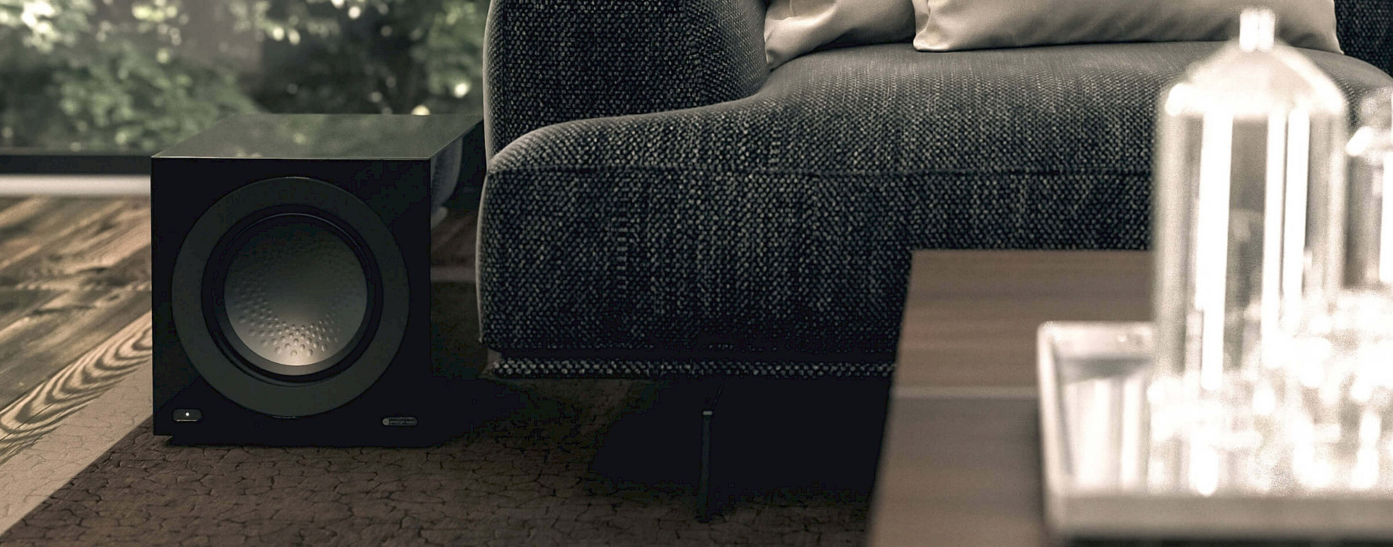 סאב וופר אקטיבי מוניטור אודיו Monitor Audio - Anthra W12 בצבע שחור מבריק מונח על רצפה צמוד לספה בסלון מודרני ומצולם בתמונת תקריב חזיתית