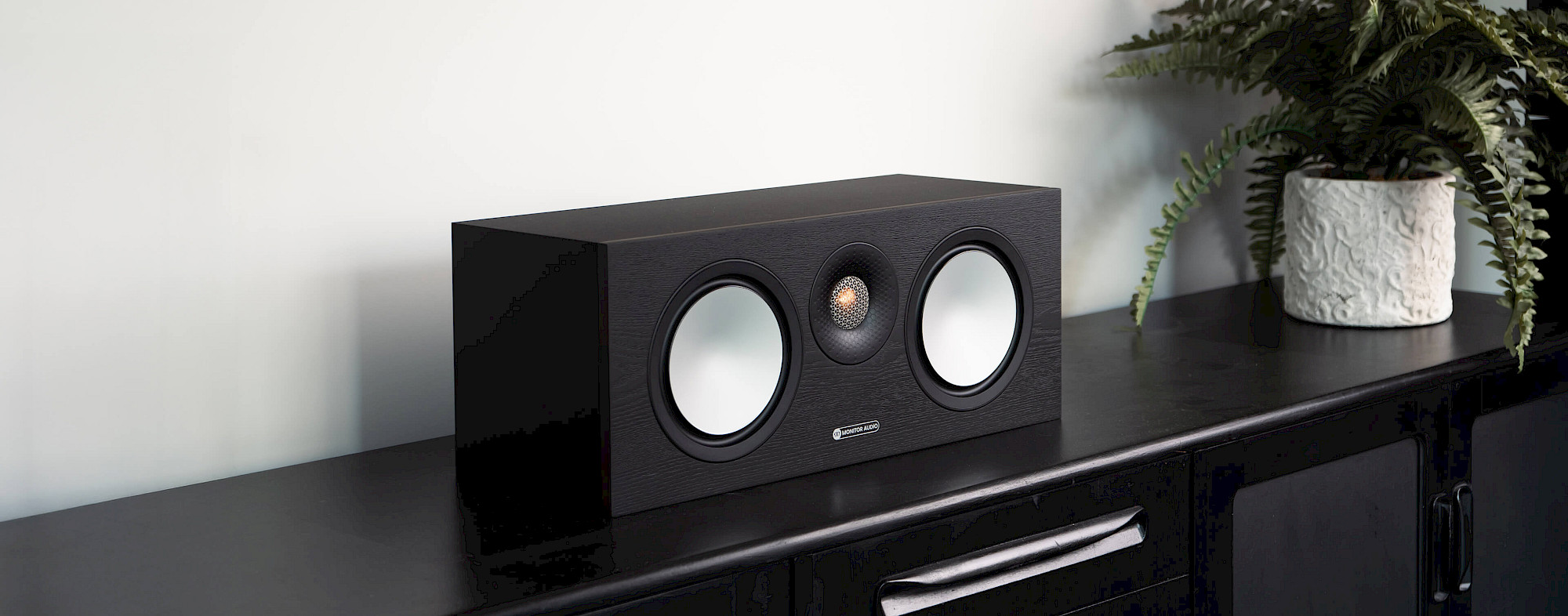 רמקול סנטר מוניטור אודיו Monitor Audio Bronze C150 7G בצבע שחור ללא גריל מונח על שידת סלון מודרנית שחורה