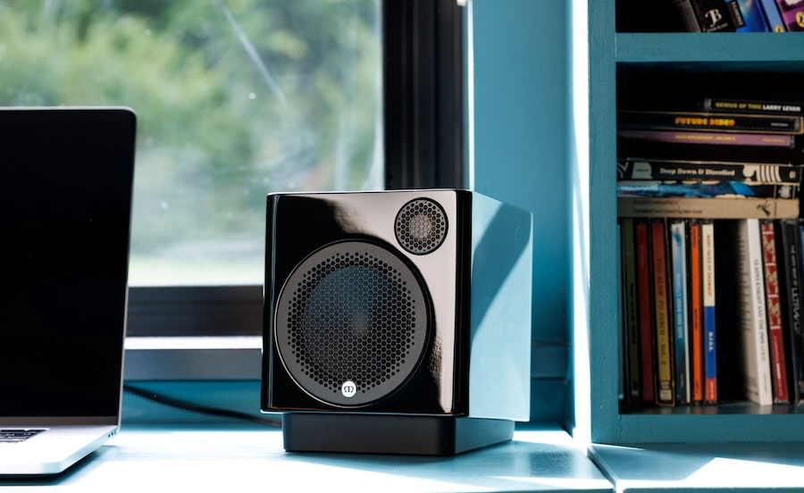 רמקול מעוצב קומפקטי מוניטור אודיו רדיוס Monitor Audio Radius 1 4G בצבע שחור מונח לצד מחשב לפטופ על שולחן עבודה משרדי