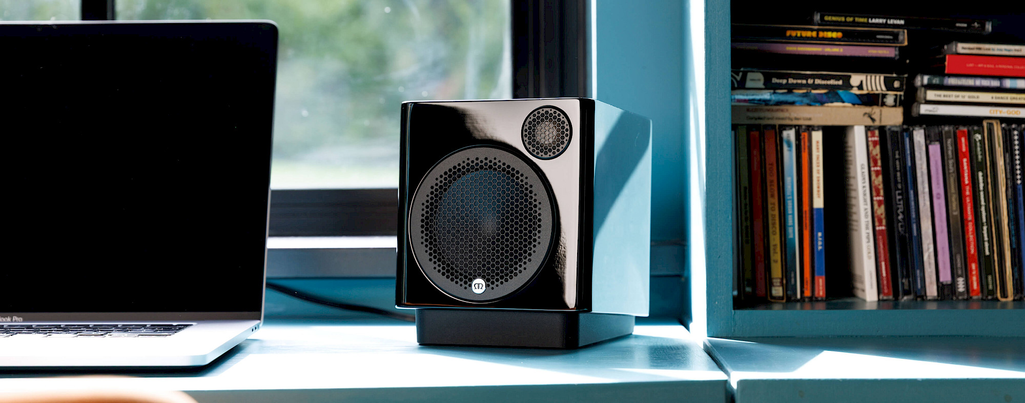רמקול מעוצב קומפקטי מוניטור אודיו רדיוס Monitor Audio Radius 1 4G בצבע שחור מונח על משטח פד יעודי בהטיילה כלפי מעלה לצד לפטופ על שולחן עבודה משרדי