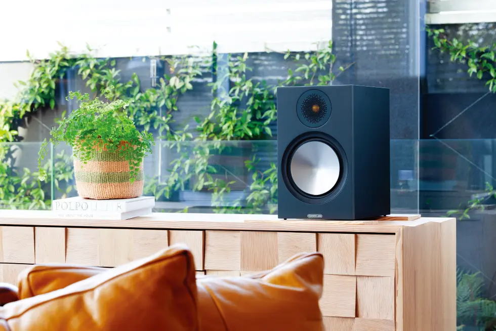 רמקול מדפי מוניטור אודיו ברונז Monitor Audio Bronze 100 6G בגימור שחור מונח על שידת עץ על רקע חלון גדול בסלון