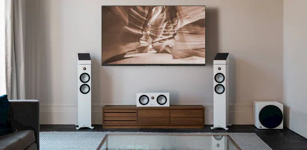 רמקולים רצפתיים מוניטור אודיו Monitor Audio Bronze 300 7G בצבע לבן עומדים בסלון משני צידי שידת טלויזיה מעץ יחד עם הסנטר, הסאב וופר וזוג רמקולי האטמוס המשלימים של אותה הסדרה