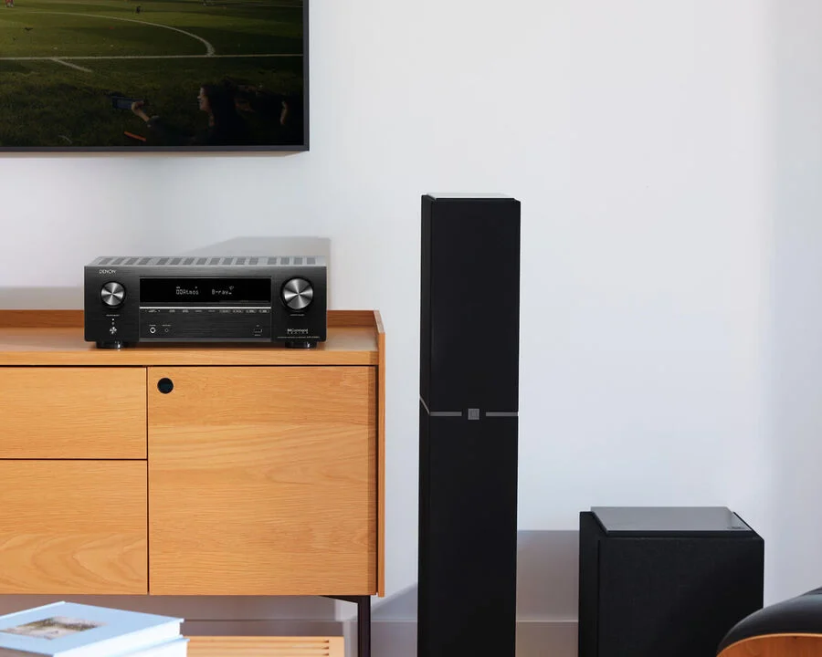 רסיבר קולנוע דנון Denon AVR-X1800H מצולם חזיתית מונח על שידת עץ מודרנית ביתית על יד רמקול רצפתי וסאב וופר אקטיבי על הרצפה מצידה הימני