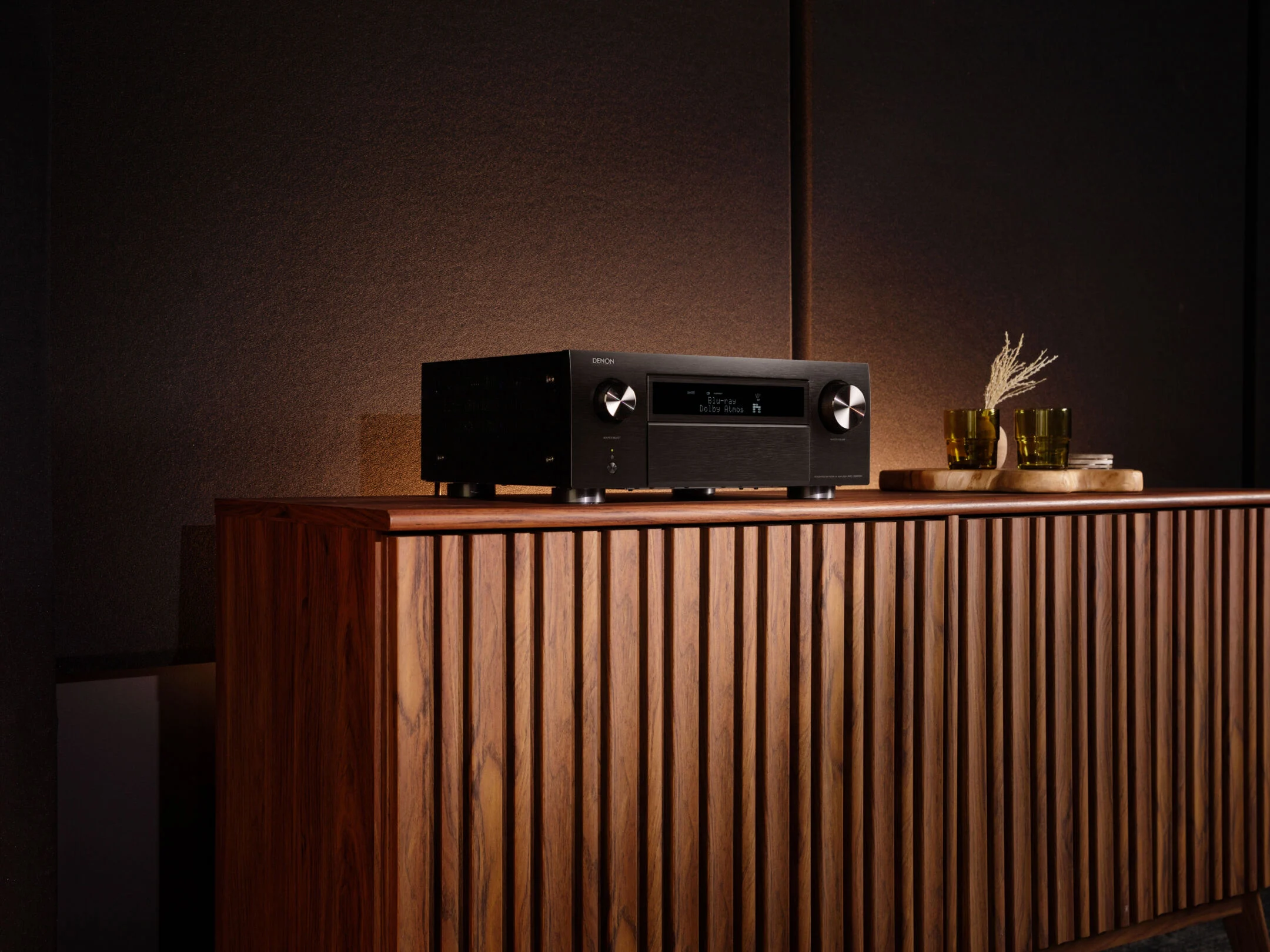 מגבר קולנוע ביתי דנון Denon AVC-X6800H עומד על שידת עץ מאסיבית בסלון מעוצב וחשוך