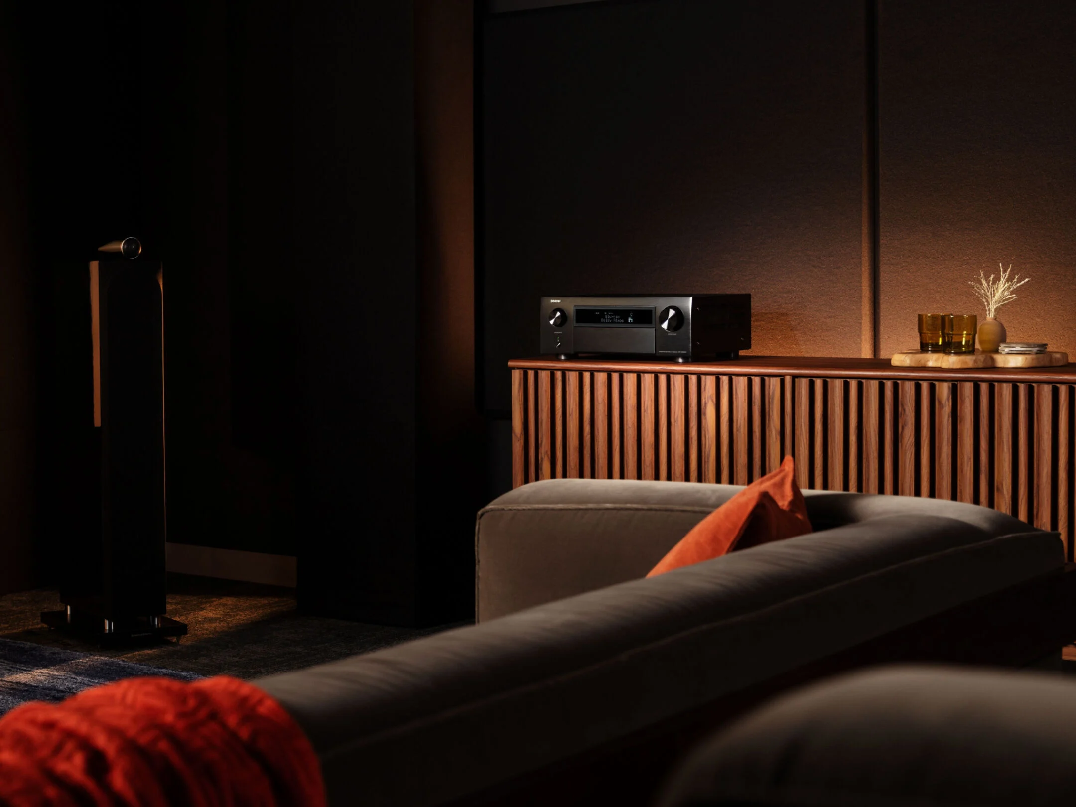 מגבר קולנוע ביתי דנון Denon AVC-X6800H עומד על שידת עץ מאסיבית בסלון מעוצב וחשוך בצילום בזווית