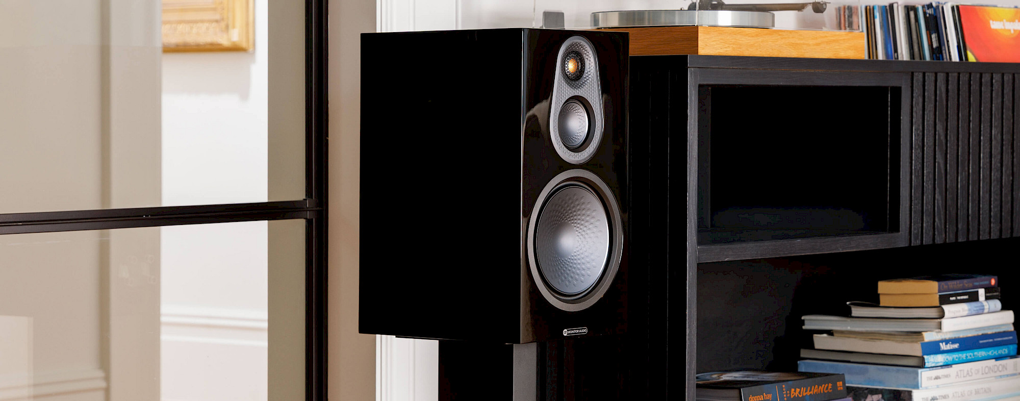 רמקולים מדפיים מוניטור אודיו Monitor Audio Silver 100 8G בצבע שחור מבריק עומדים על מעמד רצפתי לצד שידה בסלון