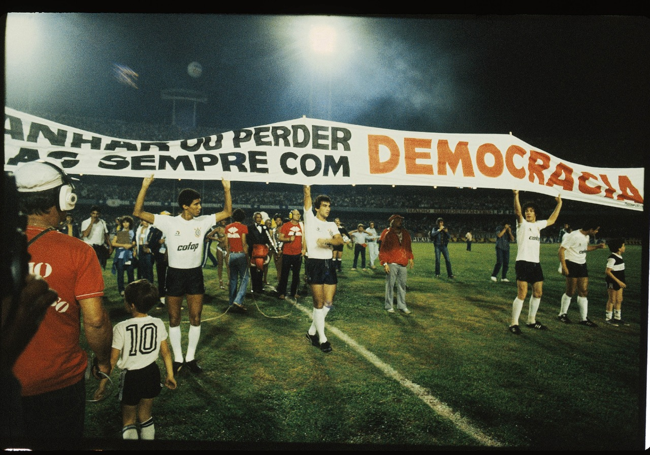 Fotografia de faixa pró-democracia em momento de transição na década de 1980, estendida por jogadores do Corinthians