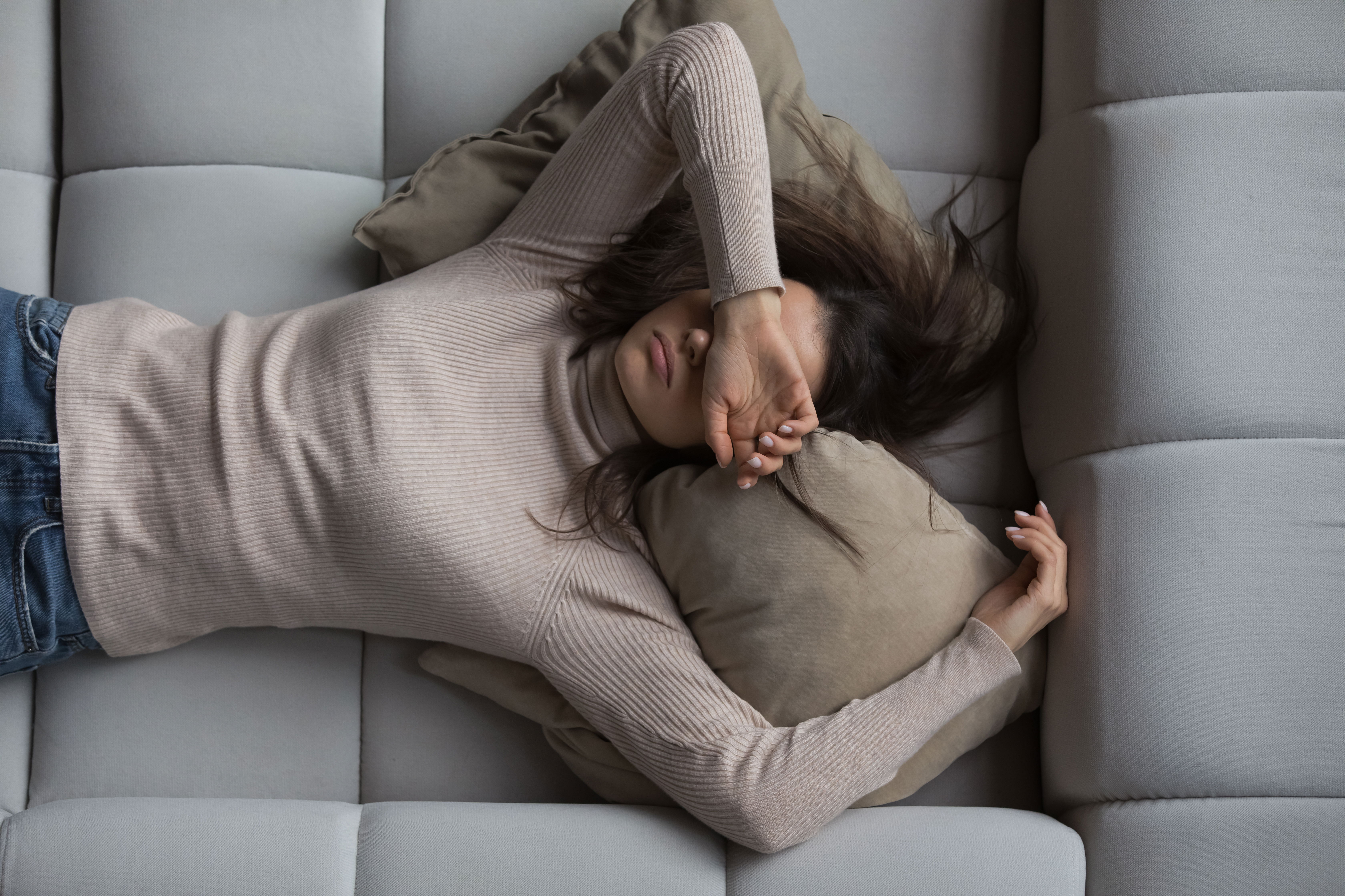 A woman lying on a couch with her arm resting over her eyes, looking fatigued. Her expression conveys exhaustion as she relaxes in a comfortable setting.