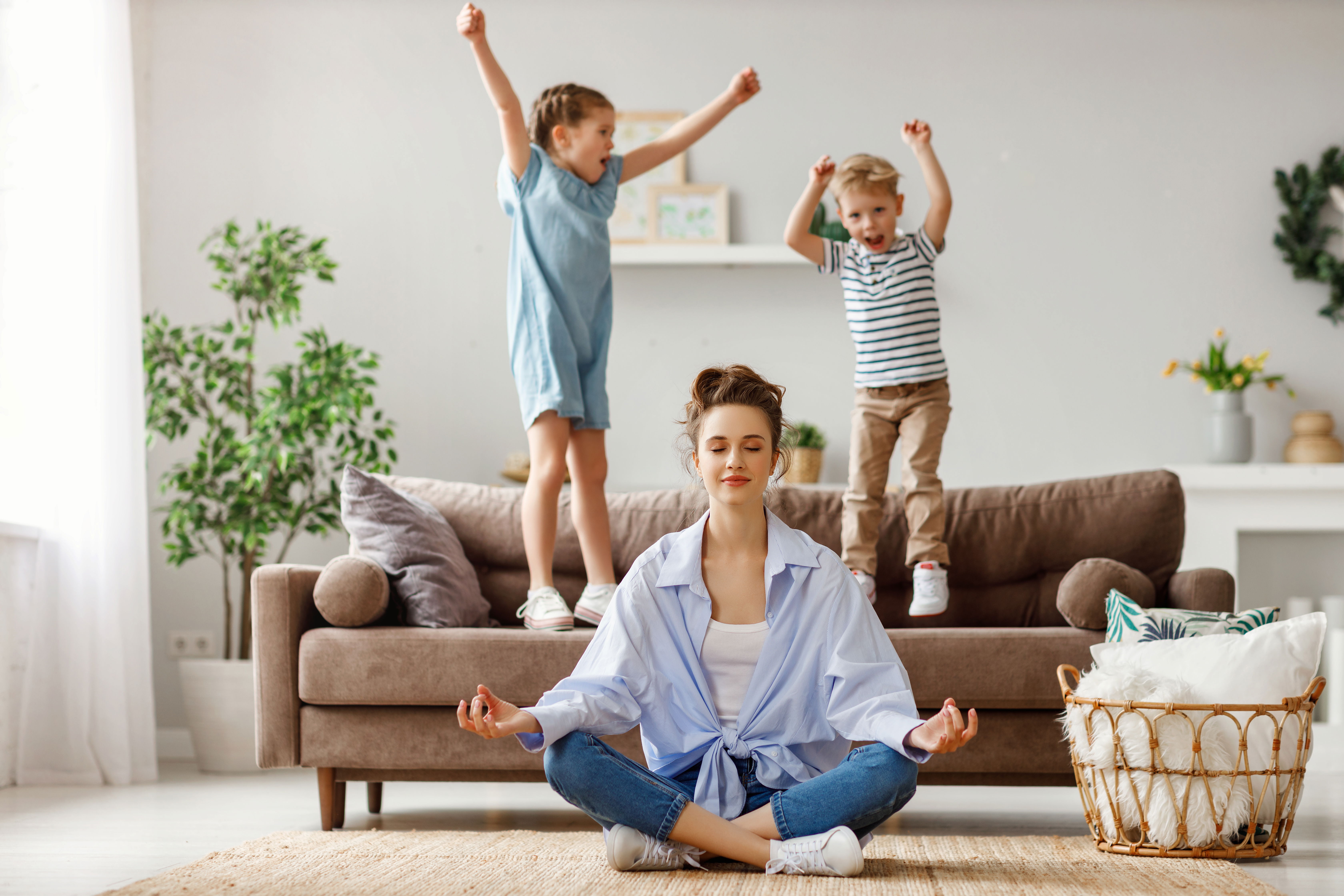  Une maman méditant en position assise jambes croisées sur le sol, pendant que ses enfants, surexcités et sautant sur le canapé, illustrent l’importance de la sophrologie pour canaliser l’énergie des enfants. 