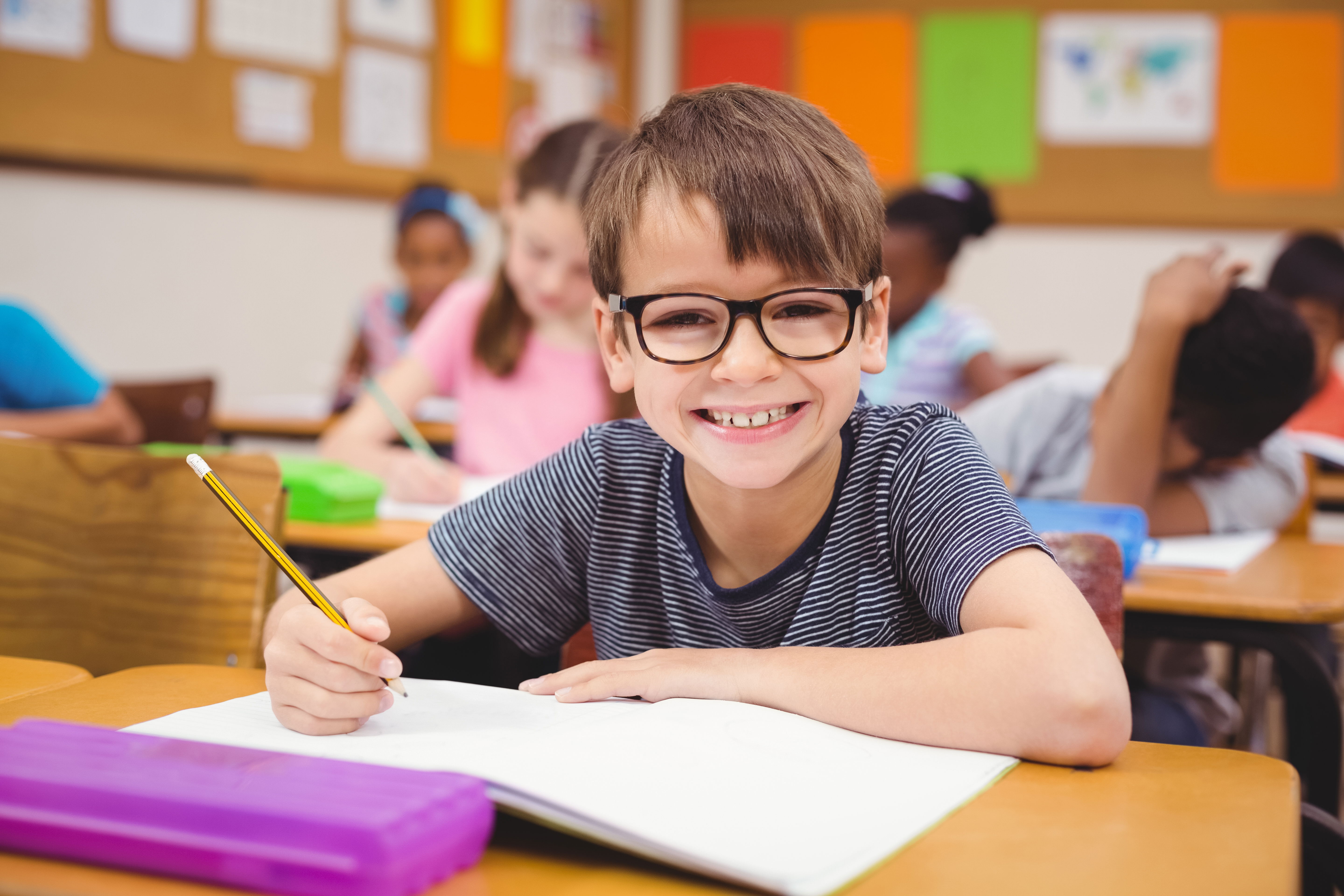 Un jeune garçon souriant dans une salle de classe, assis sur sa chaise, entouré d’autres élèves, tient un crayon, la main posée sur son cahier, illustrant la facilité d’apprentissage et la joie d’apprendre grâce à la sophrologie 