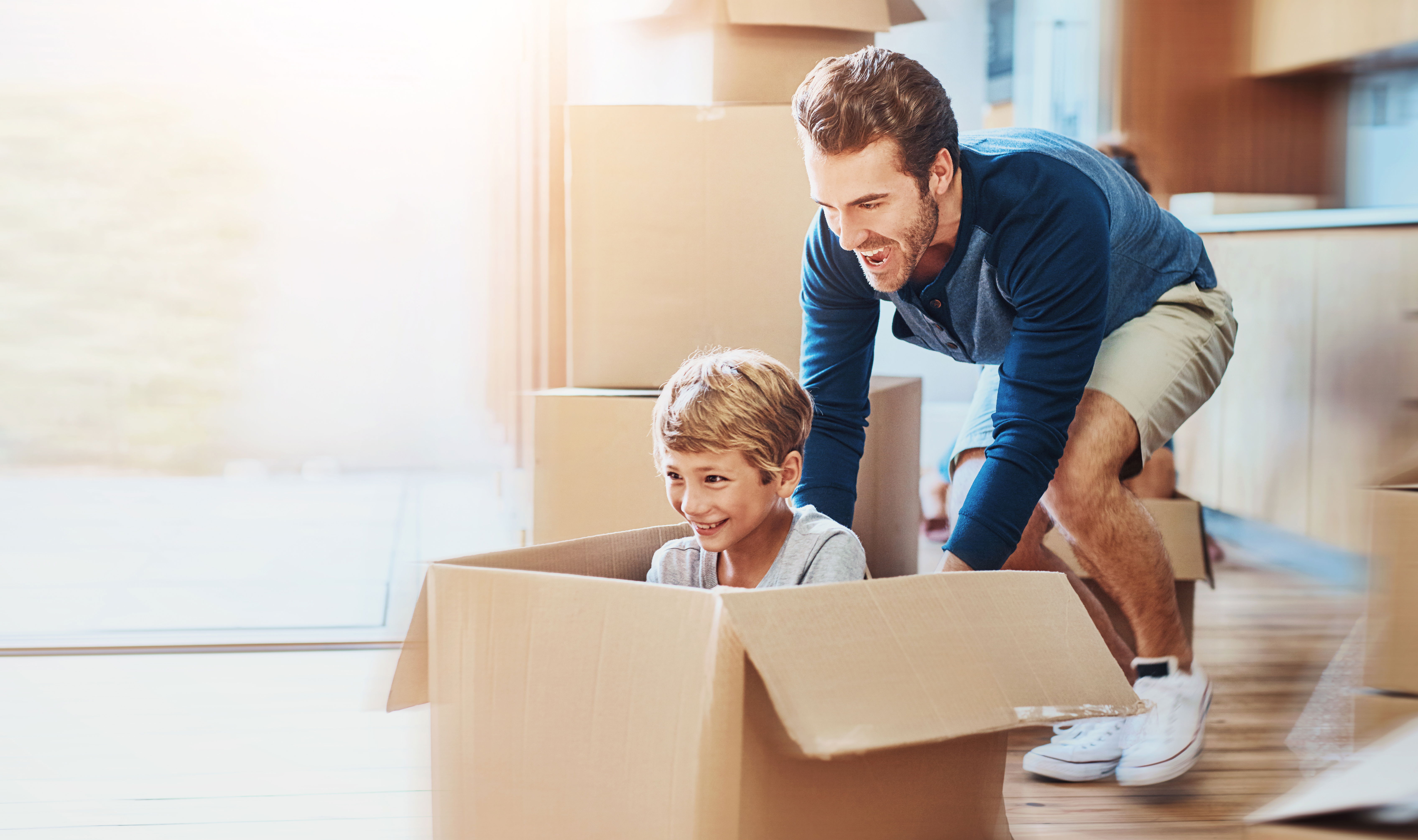 Un enfant et son père sourient et s’amusent pendant un déménagement. Le père pousse l’enfant qui est dans un carton. L’enfant est heureux. Cela illustre la facilité d’adaptation de l’enfant au changement grâce à la sophrologie. 