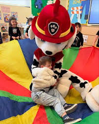 Child hugging a friendly fire dog mascot while sitting on a colorful parachute during a daycare party in Toronto