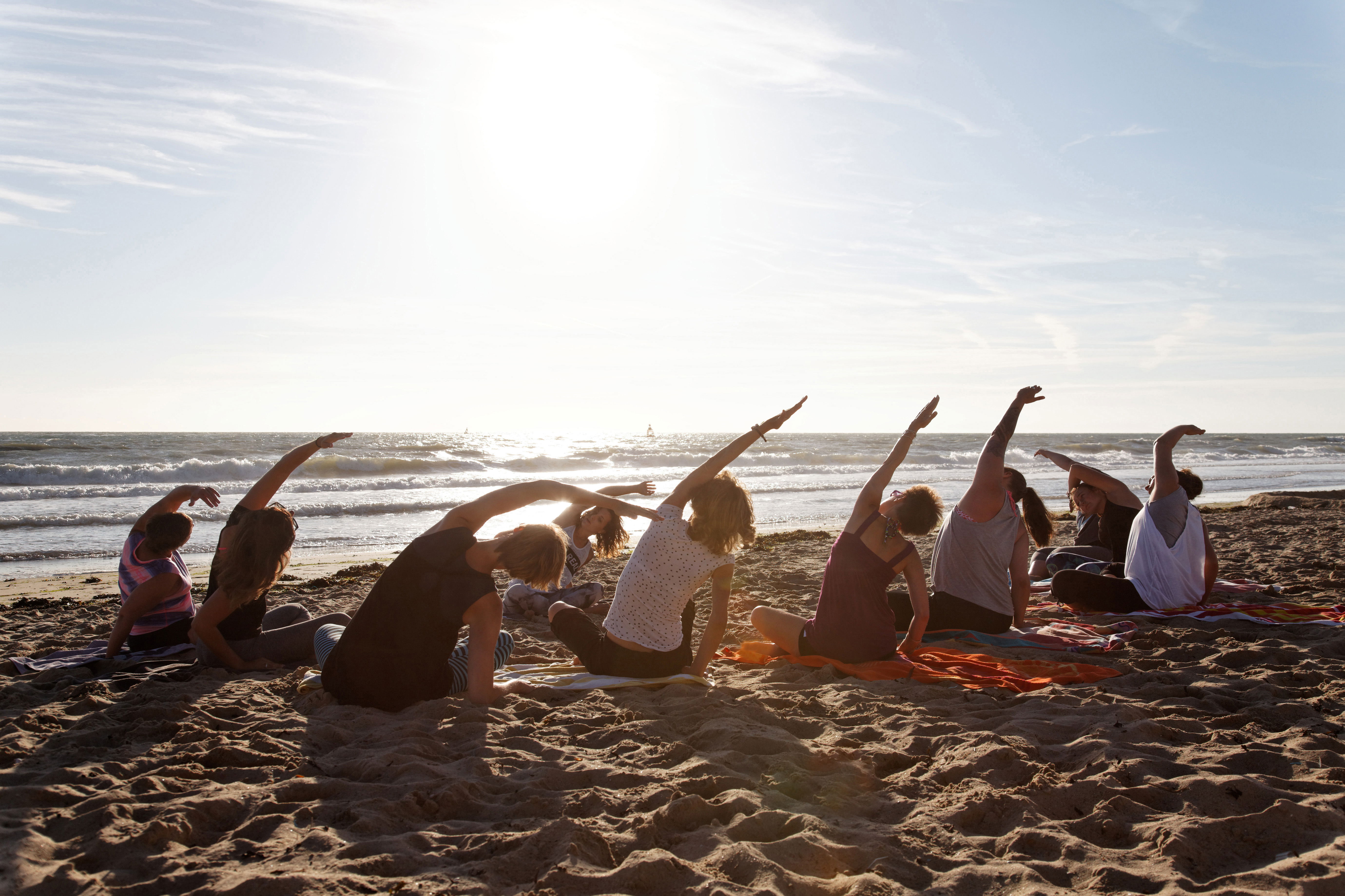 A session of sober yoga at the beach with Design to Recover sober living home in Los Angeles. A group of about 10 residents sit and do yoga under a sunset near the waterline on part of the beach near Playa Del Rey.