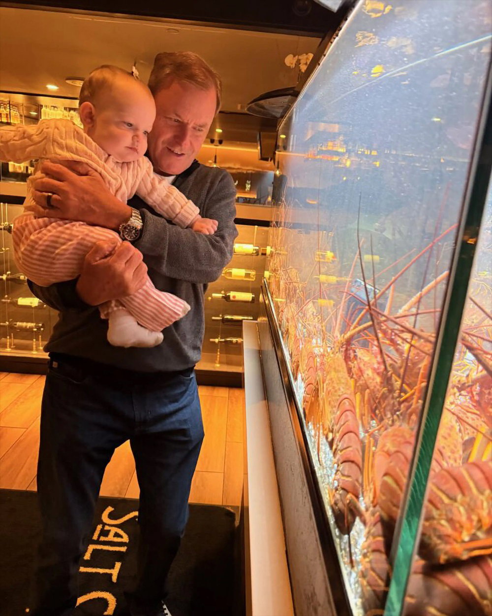 Devin Doyle holding a baby as they look closely at lobsters displayed inside a glass aquarium tank