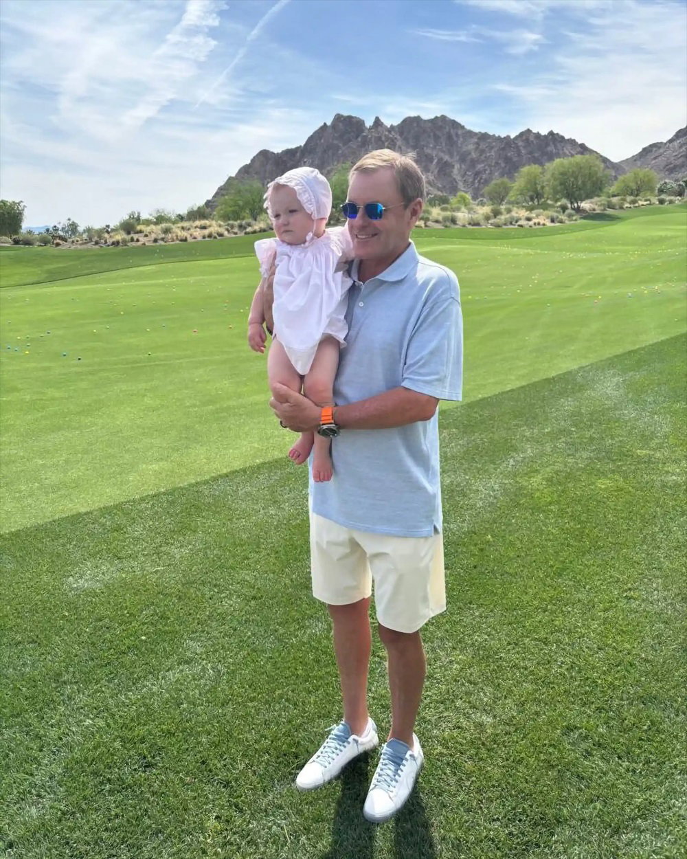 Devin Doyle smiling while holding a baby on a golf course under a bright blue sky